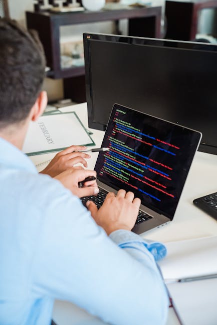 A person working at a desk with a laptop displaying lines of colorful code on the screen. The individual is wearing a light blue shirt and using a black mouse with their right hand while holding a pen