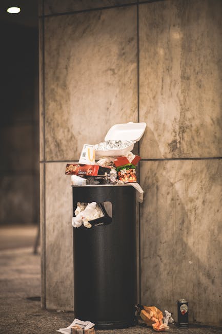 A large collection of overflowing rubbish bins and scattered waste materials on a paved urban street area, with various types of refuse including black, red, and grey wheelie bins filled with mixed ho